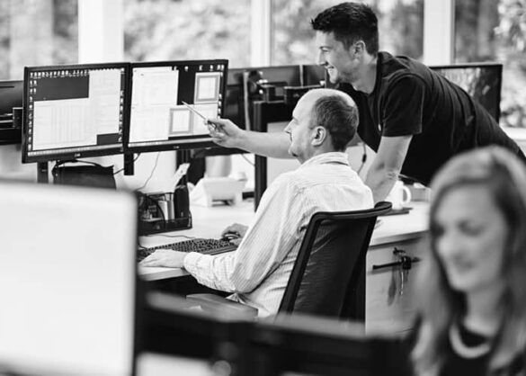 One person sat looking at a computer screen with another person stood behind, pointing at the screen.