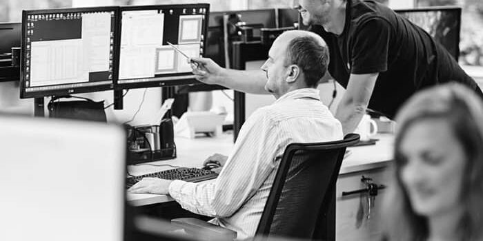 One person sat looking at a computer screen with another person stood behind, pointing at the screen.